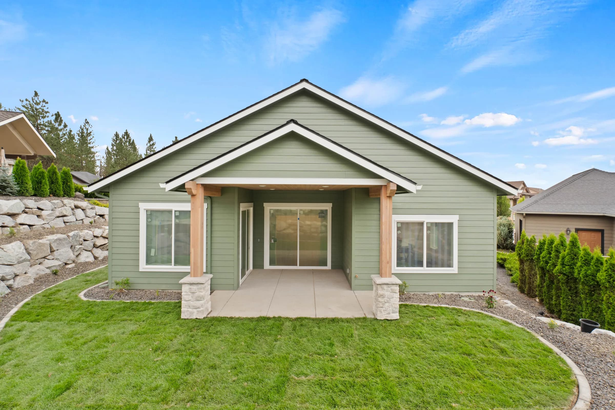 Rear of a sage-green craftsman with a timber-post covered patio and boulder terraces